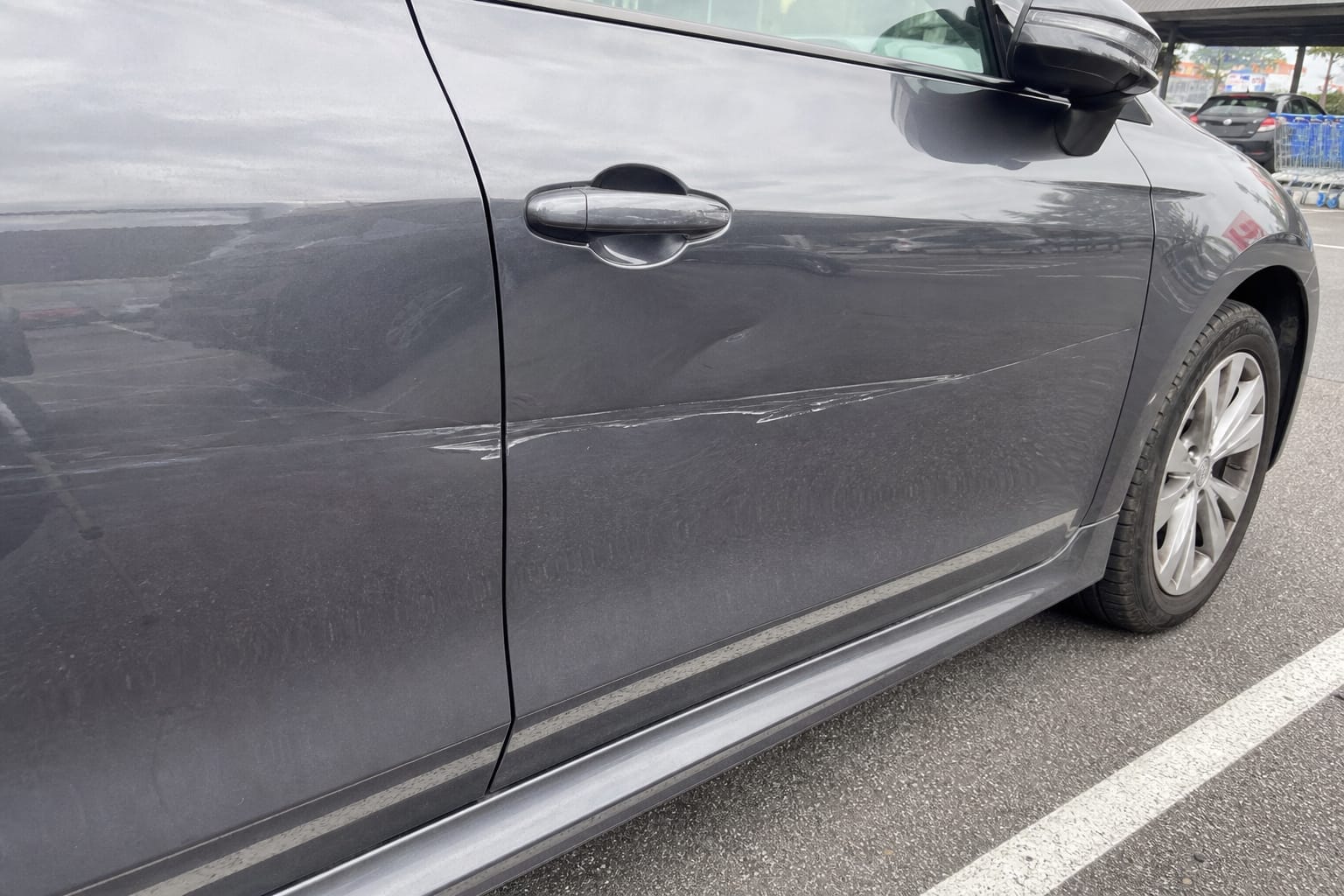 Portière de voiture rayée et légèrement enfoncée, garée sur un parking extérieur de supermarché
