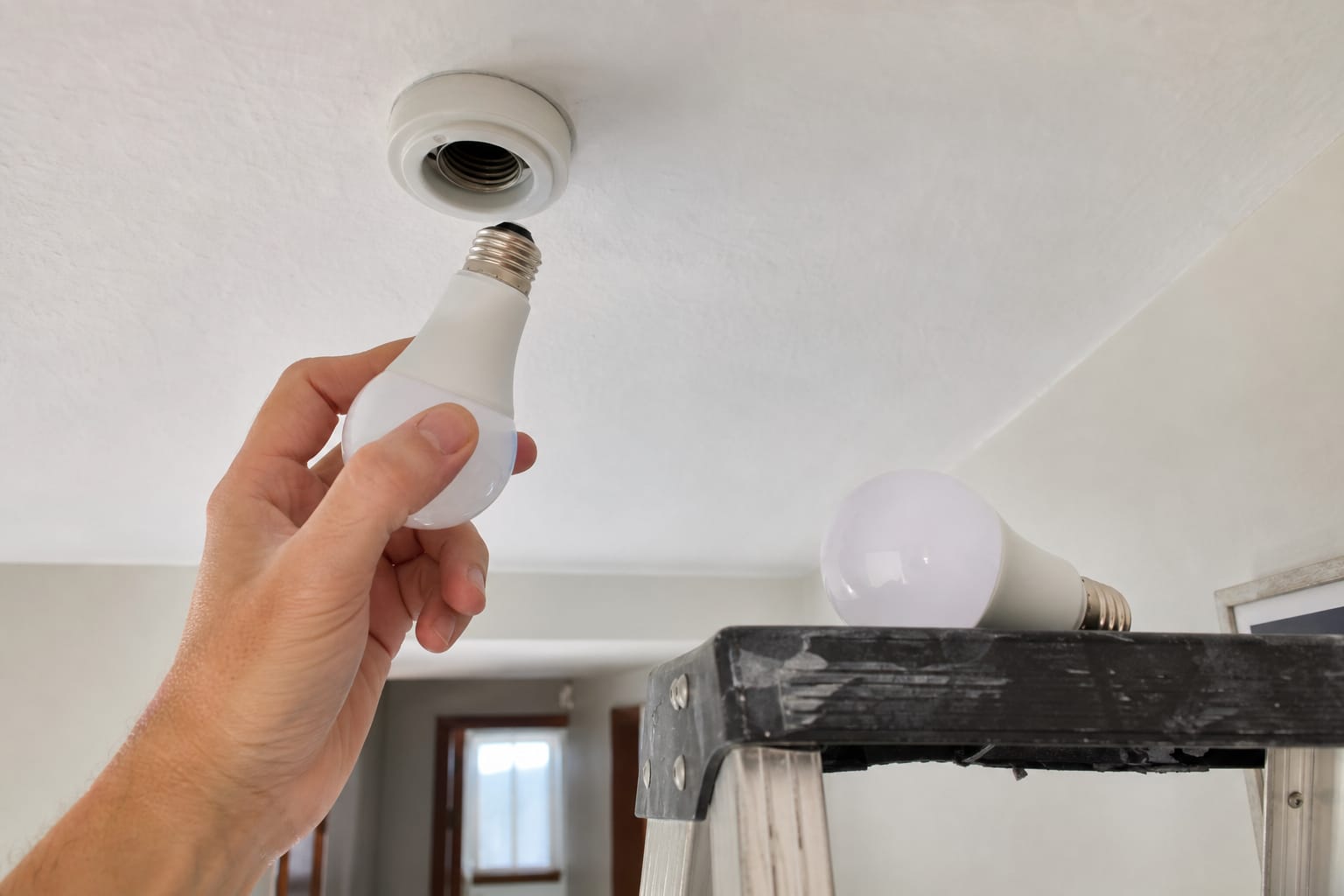 Main vissant une ampoule LED dans une douille de plafond, une autre ampoule posée sur le plateau d'un escabeau dans un couloir
